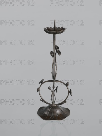 Candlestick shokudai, Takimoto craftsman, Japanese, lotus and leaf motifs, Meiji period 1868–1912, 2nd half of the 19th century, Japan, candlestick, height 44.0 cm, provenance unknown, A metal decorative stand shaped like a stylized flower with a rounded petal base, a thin stem rising from the center, a circular midsection adorned with small bud-like elements, and a lotus-shaped top plate; the object appears in dark bronze and black tones against a pale gray background.