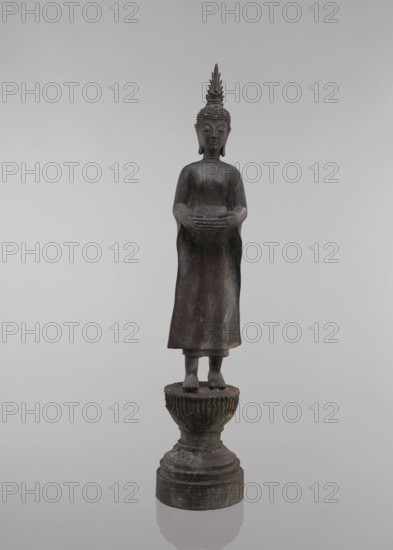 Buddha, lotus throne iconography, art of Southeast Asia, Buddha iconography, bowls, Buddhist art, 19th century, Southeast Asia, sculpture, figurine, height 54.5 cm, A standing dark bronze statue with a tall pointed headdress and textured hair, holding a rounded object with both hands at waist height, wearing a long robe that falls to the ankles; the figure stands barefoot on a carved pedestal featuring a fluted bowl shape atop a stepped circular base. The visible colors are dark brown, muted greenish patina, and hints of reddish-brown against a neutral gray background.