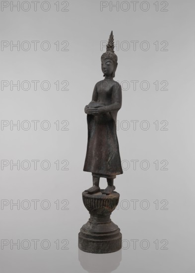 Buddha, lotus throne iconography, art of Southeast Asia, Buddha iconography, bowls, Buddhist art, 19th century, Southeast Asia, sculpture, figurine, height 54.5 cm, A standing dark brown bronze statue with a greenish patina shows a robed figure on a round tiered pedestal; the figure wears a tall pointed headdress with textured hair and holds a rounded object in both hands at waist level, with a long skirt-like garment draping to the ankles and bare feet positioned on a fluted cushion-like support, all set against a pale gray background.