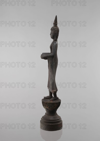Buddha, lotus throne iconography, art of Southeast Asia, Buddha iconography, bowls, Buddhist art, 19th century, Southeast Asia, sculpture, figurine, height 54.5 cm, A standing bronze statue on a pedestal showing a slender humanlike figure in profile, with textured hair forming a pointed topknot, elongated earlobes, and a smooth face and torso; the figure wears a long draped garment that falls to the ankles and holds a shallow bowl with both hands at waist level, while both feet rest on a decorative round platform atop a stepped circular base; visible colors include dark brown and greenish patina on the metal with hints of reddish-brown, set against a neutral pale gray background.