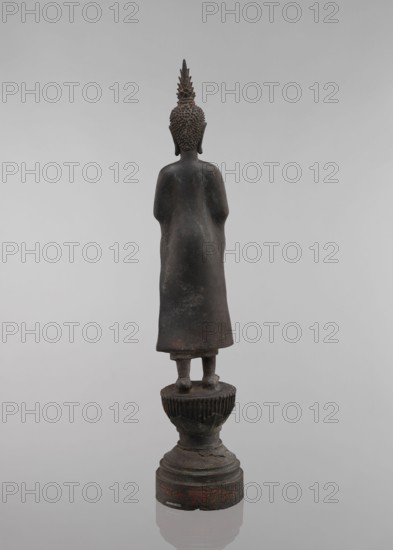 Buddha, lotus throne iconography, art of Southeast Asia, Buddha iconography, bowls, Buddhist art, 19th century, Southeast Asia, sculpture, figurine, height 54.5 cm, A dark bronze statue of a standing figure viewed from the back on a tiered pedestal, the figure wearing a long robe with smooth contours and a textured hair or headpiece ending in a pointed finial; the pedestal shows carved fluting and rounded stacked forms with some surface irregularities and wear. The visible colors are dark brown, black, and hints of reddish-brown on the base, set against a pale gray background.