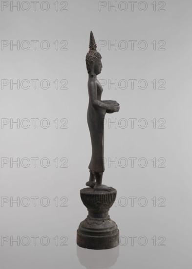 Buddha, lotus throne iconography, art of Southeast Asia, Buddha iconography, bowls, Buddhist art, 19th century, Southeast Asia, sculpture, figurine, height 54.5 cm, A tall dark bronze statue of a standing figure shown in profile on a round tiered base, with a textured conical headdress and a smooth elongated robe; the figure holds a shallow bowl with both hands at waist height and stands with feet together on a decorative pedestal, against a plain light gray background with shades of darker gray on the statue and base.