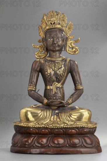 Amitayus, Buddhist/Tibetan/Himalayan/Mongolian art; Himalayan and Mongolian culture; Buddha iconography with ushnisha, lotus throne, vajras; sculpture/figurine/cult object; inner Mongolia; 2nd half of the 18th–1st half of the 19th century; height 37.5 cm; weight 2.12 kg., A seated sculptural figure with a dark brown body and bright gold decorative elements, wearing an ornate gold crown with floral and scrolling motifs and gold ear ornaments; the figure has a calm facial expression and a small circular mark on the forehead, with gold detailing on the chest and a sash across the torso; the arms and hands rest in the lap with gold bracelets and arm ornaments, and the lower body is draped in gold-adorned garments; the figure sits on a carved wooden pedestal with a reddish-brown finish and rounded lotus petal shapes; the background is a plain neutral gray.