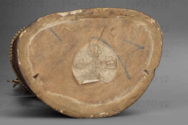 Amitayus; creator, Buddhist/Tibetan/Himalayan/Mongolian art; Himalayan and Mongolian cultural origins; Buddha iconography with ushnisha; lotus throne; vajras; 2nd half of the 18th–1st half of the 19th century; Inner Mongolia; sculpture figurine, cult object; height 37.5 cm; weight 2.12 kg., The image shows the underside of an object with a worn wooden base in shades of tan and light brown, bordered by a rim of darker beige and pale cream where the material is chipped; a faded circular paper label with a scalloped edge and brownish line drawings is affixed near the center with a small curled lift at its edge; several short blue marks are visible on the wood surface; along the left edge a row of small rounded metallic beads in a dull gold color peeks into view, and a small dark brown triangular wood fragment is attached near the left interior; the background is a smooth medium gray.