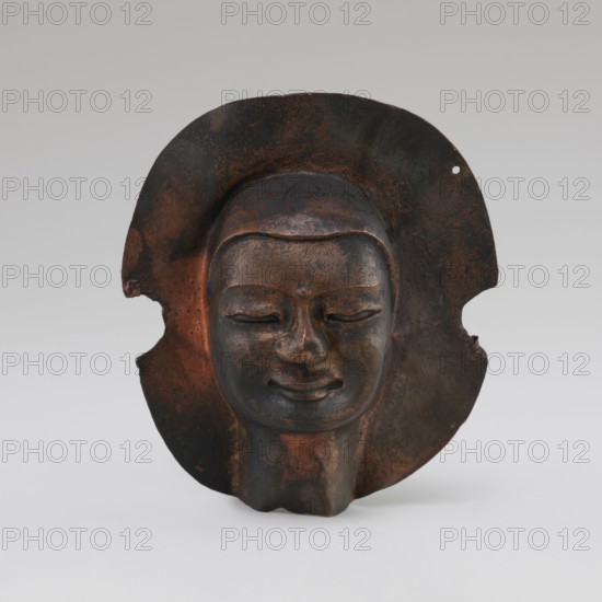 Head, unknown provenance; Tibetan culture; monks, disciples; Tibetan Buddhist art, Buddhist art; faces; 19th–20th century; Tibet place of origin?; figurine sculpture; height 8.9 cm, width 3.5 cm, A sculpted face mask with a smooth, rounded forehead and closed eyes set within a broad, irregular halo-like backplate, showing a serene, slightly smiling mouth and a rounded chin; the surface displays a mix of dark brown, rusty orange, and black patina with patches of lighter brown and subtle metallic sheen, and the backplate has irregular edges and a small perforation on one side with areas of wear and mottled coloration.