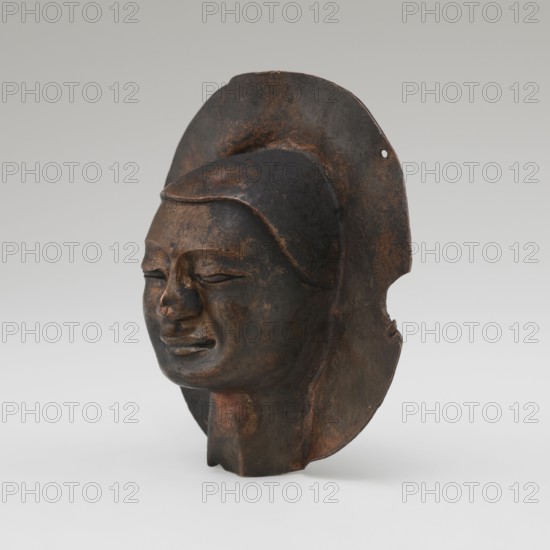 Head, Tibetan, monks, disciples, Tibetan Buddhist art, Buddhist art, faces, 19th/20th century, Tibet place of origin ?, figurine sculpture, height 8.9 cm, width 3.5 cm, A sculpted head of dark brown and black tones with areas of rusty brown highlights, showing a serene facial expression with closed eyes and a subtle smile; the surface appears worn and textured, with a rounded headdress or halo-like backing that extends behind and around the head, and a short neck base supporting the piece.