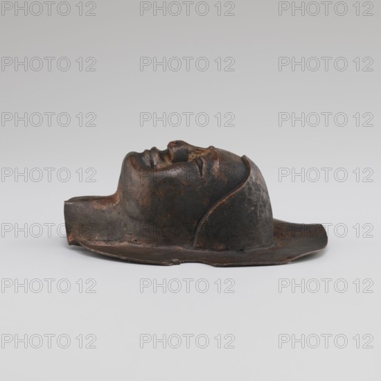 Head, unknown provenance; Tibetan culture; monks, disciples; Tibetan Buddhist art, Buddhist art; faces; 19th–20th century; Tibet place of origin ?; figurine sculpture; height 8.9 cm, width 3.5 cm., A dark brown and black sculpted object resembling a reclining human head with closed eyes rests on a flat base; the surface shows variations of brown, rust, and black tones with a slightly rough, mottled texture, and the form includes curved facial contours, a prominent nose, lips, and an outline suggesting hair or a cap, all set against a light gray background.