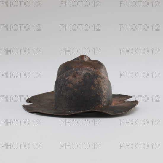 Head, unknown provenance; Tibetan; monks, disciples; Tibetan Buddhist art, Buddhist art; faces; 19th/20th century; Tibet place of origin?; figurine sculpture; height 8.9 cm, width 3.5 cm, A worn metal hat with an irregular brim and a domed crown sits on a neutral surface; the object shows a mottled surface with shades of dark brown, rusty orange, and black, with areas of uneven texture and small patches of lighter brown.