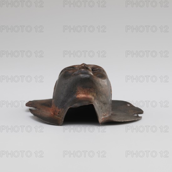 Head, Tibetan, monks, disciples, Tibetan Buddhist art, Buddhist art, faces, 19th/20th century, Tibet, figurine, sculpture; height 8.9 cm, width 3.5 cm, A sculpted metallic face mask lying on a flat surface, viewed from the front with the chin nearest the camera, showing closed eyes, a nose, and relaxed mouth features; the mask has an irregular skirt-like flange around its lower edge with uneven, slightly torn contours and small perforations, and the surface displays a mottled patina with dark brown, black, copper, and hints of reddish tones against a pale gray background.