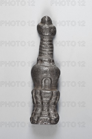 Thogcha in the shape of a stupa chorten, creator, Tibetan Buddhist/Himalayan art; amulet-shaped sculpture; material unspecified; function: cult object; subject: stupa; provenance, date: 18th–1st half of 20th century ; place of origin: Tibet; dimensions: height 5.6 cm, width 1.6 cm., A vertically oriented dark metallic object with a textured surface sits against a light gray background; its top tapers into a rounded head, the midsection narrows into a ribbed neck, the central portion broadens into a smooth, bulbous form with a shallow indentation, and the lower section flares into a carved, grid-like base with raised, blocky details. The visible colors are dark gray and light gray.