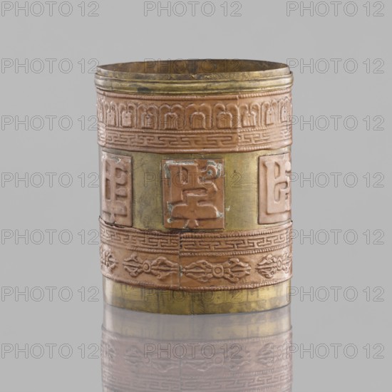Cylinder; creator, craft; Tibetan; 19th century; Mongolia and Tibet place of origin; applied copper sheet; inscriptions; vajras; length 7.3 cm; diameter 6.0 cm., A cylindrical metal container with decorative horizontal bands, showing a mix of coppery brown and muted gold tones, stands against a neutral gray background with a subtle reflection below; the container has a textured rim and several embossed rectangular panels and ornamental patterns in coppery brown, separated by gold-colored vertical strips, and a few areas show darker tarnish or wear.