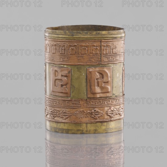 Cylinder, craft, Tibetan, inscriptions, vajras, applied copper sheet, 19th century, Mongolia, Tibet, varia, length 7.3 cm, diameter 6.0 cm, A cylindrical metal container with decorative raised bands and panels, shown against a neutral gray background and reflected faintly on a glossy surface beneath; the object displays a mix of metallic colors including dull gold, copper, and brass tones, with areas of patina and darker spots, and features ornamental motifs in relief such as repeating geometric borders, stylized floral or rope-like patterns, and several raised square panels with abstract shapes, the top rim being slightly uneven and the interior appearing darker than the exterior.