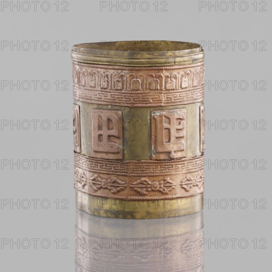 Cylinder, craft, Tibetan, inscriptions, vajras, applied copper sheet, 19th century, Mongolia, Tibet, varia, length 7.3 cm, diameter 6.0 cm, A cylindrical metal vessel with decorative raised bands and panels sits against a light gray background. The vessel shows a mix of metallic colors including brass yellow, coppery pink, and hints of greenish patina. The top edge is slightly uneven and darker brass in color, followed by a raised coppery band with repeating arched motifs. Below that is a broader section with alternating square raised panels and flat vertical strips, the panels in coppery pink and the strips showing brass yellow with areas of greenish discoloration. A narrow decorative band with a geometric pattern separates this from a lower coppery band featuring floral or foliate relief motifs, and the base returns to a brass yellow tone with wear and slight tarnish. The vessel rests on a reflective surface that produces a soft mirror image of the lower part.