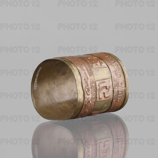Cylinder, craft, Tibetan culture, inscriptions, vajras, applied copper sheet, 19th century, Mongolia and Tibet place of origin, varia, length 7.3 cm, diameter 6.0 cm, A cylindrical metal object lying on its side with a reflective surface beneath it, showing a mirrored image; the cylinder has bands of different textures and raised rectangular panels around its exterior, with decorative floral and geometric patterns; the inner surface appears smooth and slightly worn; visible colors include brassy yellow, coppery brown, pale gold, and muted gray in the background and reflection.