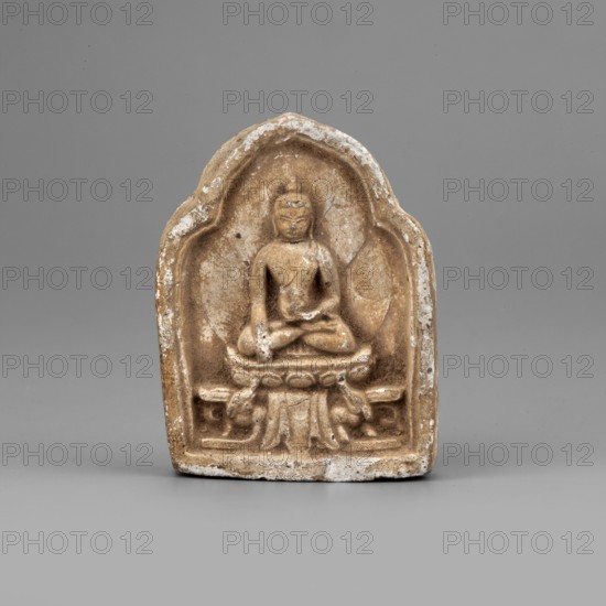 Caca Tib. tsa-tsa depicting Shakyamuni Buddha; creator, Buddhist/Tibetan/Himalayan art; iconography: Buddha, lotus throne, ushnisha, mudras; 19th–1st half of 20th century; possible origin: Mongolia or Tibet; sculpture/figurine/cult object; dimensions: height 5.1 cm, width 3.9 cm, depth 1.25 cm., A small carved object in beige and light brown tones sits against a smooth gray background; the object has a rounded arch shape with a recessed central area containing a seated figure in a meditative pose on a raised platform, with carved folds and decorative elements beneath the platform and a textured surface showing lighter worn spots and white residue along the edges.