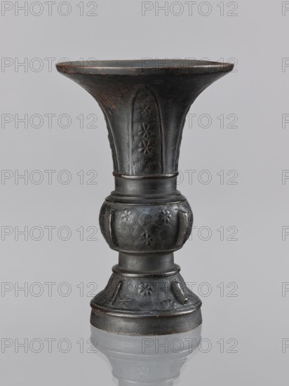 Gu-type vase gu cup, gu cups, Chinese art, Chinese culture, ceremonial ritual vessel with spiral and plant motifs, Qing dynasty 1644–1911, 18th century, China place of origin ?, vessel, height 14.0 cm, diameter 10.0 cm, A dark-colored metal vessel with a flared rim and a pedestal base, showing a patina with brownish wear at the rim and edges; the neck flares outward and is decorated with embossed swirling and floral patterns set within vertical panels; below the neck a rounded bulbous section bears similar embossed swirls and floral motifs, leading to a narrower cylindrical segment and a flared base that repeats the embossed decorative panels; the surface appears predominantly dark gray to black with areas of brownish rust and lighter gray highlights, and the object sits on a reflective pale gray surface against a neutral light gray background.