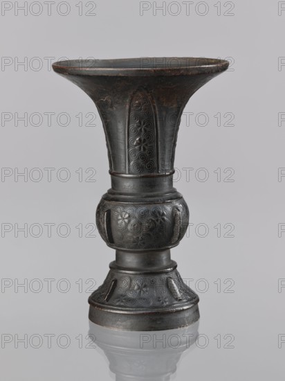 Gu-type vase, gu cup, Chinese, ceremonial/ritual vessel, spiral and plant motifs, Qing dynasty 1644–1911, 18th century, China place of origin ?, height 14.0 cm, diameter 10.0 cm, A dark metallic vase-like object with a flared rim, a narrow waist, and a rounded midsection, standing against a light gray background; the surface shows embossed circular and spiral patterns and subtle floral motifs; the metal appears dark gray to black with areas of brownish wear and slight greenish patina; the object casts a soft reflection on a glossy surface below.