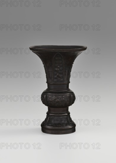 Gu-type vessel, gu cups, Chinese art, Chinese culture, ceremonial vessels, Qing 1644–1911, ritual vessels, 18th century, China, vessel, height 15 cm, diameter 10.7 cm, A dark bronze vessel with a flared rim stands upright against a neutral gray gradient background; the vessel has a wide, shallow bowl that tapers into a decorated neck and a bulbous midsection with raised ornamental motifs, followed by a narrower cylindrical stem and a flared round foot with carved swirling patterns. The visible colors are dark brown to near-black on the vessel and various shades of gray in the background and surface beneath it.
