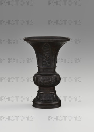 Gu-type vessel gu cup, Chinese, Qing dynasty 1644–1911, 18th century, ritual/ceremonial vessel, height 15 cm, diameter 10.7 cm., A dark bronze vessel with a wide flared rim tapering into a narrow waist and then expanding into a rounded midsection before narrowing again to a decorated base, standing against a neutral gray gradient background. The surface of the vessel shows intricate raised decorative motifs in repeated swirling patterns and stylized animal or mythical creature figures on the midsection and upper body, with carved vertical panels on the flared portion. The overall color palette is dark brown to nearly black with subtle lighter brown highlights on raised areas, and the background is shades of gray.