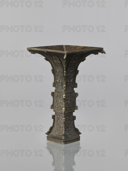Fanggu vase, fanggu vessel, Chinese art, Chinese culture, ceremonial/ritual vessel, gu cup form, decorated with beasts fantasy, birds, dragons iconography, Qing dynasty 1644–1911, 19th century, China; vessel; height 19.9 cm, width 11.5 cm, depth 11.8 cm., A tall, narrow metal vessel with a flared square rim and a stepped square base stands centered against a pale gray background; the object shows a weathered dark bronze surface with greenish patina and small accents of brighter, golden metal at corner fittings, and its vertical body is divided into stacked sections decorated with raised, curvilinear relief patterns and flared ornamental projections, while the top interior appears darker and the base casts a soft reflection on the glossy surface beneath.