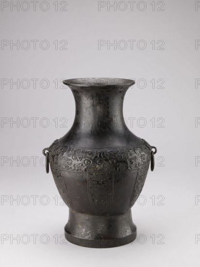 Hu-type vase, Chinese, ceremonial ritual vessel with handles and taotie iconography, Qing dynasty 1644–1911, 19th century, China; vessel; height 39 cm, width 27.5 cm, diameter 24.5 cm., A single dark metal vase with a flared rim and a narrow neck that widens into a rounded shoulder and tapers down to a short pedestal base, the surface showing decorative raised patterns around the upper body and subtle textured wear, two small looped handles on opposite sides each holding a ring, and visible colors including dark gray, black, and hints of brown against a light neutral background.