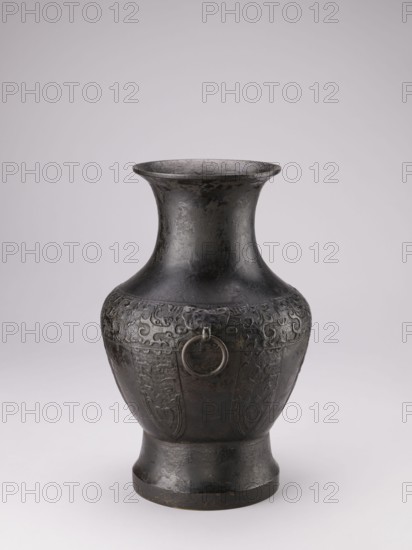 Hu-type vase, Chinese, ceremonial ritual vessel with handles and taotie iconography, Qing dynasty 1644–1911, 19th century, China; vessel, height 39 cm, width 27.5 cm, diameter 24.5 cm., A single dark metal vase stands against a pale background, showing a rounded body that tapers to a flared neck and sits on a slightly stepped base. The surface of the vase is textured and shows areas of wear and subtle variations in dark gray and black tones with hints of brownish patina. Around the upper portion of the body there is a band of low-relief decorative motifs and a few raised panels with carved designs, and a ring handle set into a small sculpted mount is visible on one side. The overall shape is symmetrical and the lighting creates soft highlights and gentle shadows that emphasize the vessel’s curved profile and detailed surface.