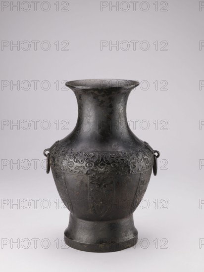 Hu-type vase, Chinese, ceremonial ritual vessel with handles and taotie iconography, Qing dynasty 1644–1911, 19th century, China; vessel, height 39 cm, width 27.5 cm, diameter 24.5 cm., A dark metal vase with a flared rim and a narrow neck that widens into a bulbous body, standing on a slightly flared foot; the surface shows an overall dark gray to black patina with areas of lighter gray wear and subtle brownish spots; the upper portion of the body features a band of raised ornamental patterns and a few ring handles attached on opposite sides, while the lower body has vertical panel-like decorations in low relief; the background is a smooth, pale gray.
