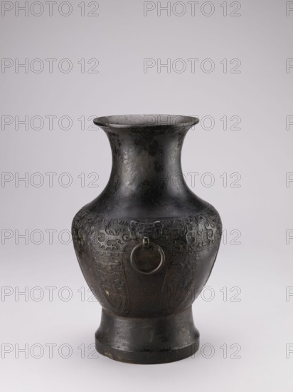 Hu-type vase, Chinese, ceremonial vessel with handles and taotie iconography, ritual vessel, Qing dynasty 1644–1911, 19th century, China; vessel; height 39 cm, width 27.5 cm, diameter 24.5 cm., A dark metal vase with a flared rim and a narrow neck widening into a rounded body set on a slightly flared foot, the surface showing a worn, textured patina in shades of black and dark gray with areas of lighter gray and subtle brownish tones, decorative low-relief patterns circling the upper body that include flowing, scroll-like motifs, and a metal ring attached to the body through a small projecting element, all set against a pale neutral background.