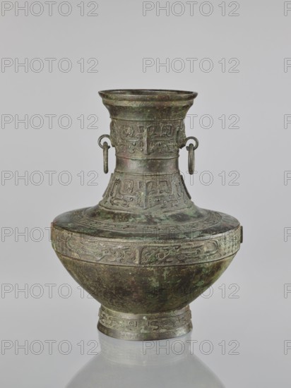 Hu-type vessel, Chinese, ceremonial ritual vessel, Qing dynasty 1644–1911, 19th century, Chinese origin, hu shape with handles, decorated with fantastical beasts, height 33.5 cm, diameter 27.0 cm., An ornate metal vessel with a wide rounded body, a narrow foot, and a tall flaring neck, showing a mottled surface of dark green, brown, and hints of reddish patina; the vessel is decorated with raised carved bands of repeating abstract motifs and geometric patterns around the neck, shoulder, and foot, and has a pair of small looped handles with attached rings on opposite sides of the neck; the object is photographed against a plain light gray background with a faint reflection visible beneath it.