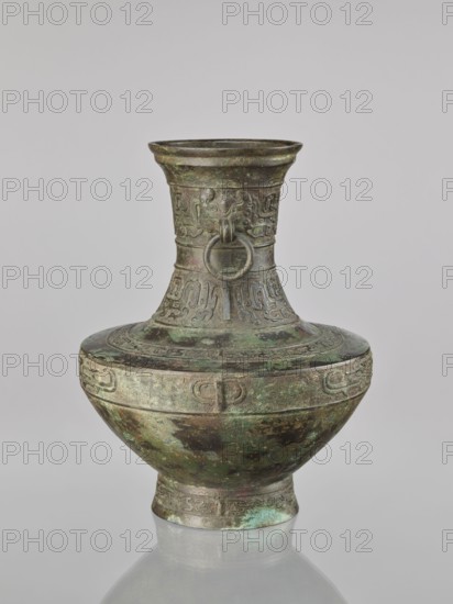 Hu-type vessel, Chinese, ceremonial/ritual vessel, Qing dynasty 1644–1911, possibly 19th century, decorated with fantastical beasts, with handles; vessel, height 33.5 cm, diameter 27.0 cm., A single metal vessel with a rounded body, narrow neck, flared rim, and short foot is shown against a neutral background; the vessel has a patinated surface with visible green, brown, dark green, and mottled black tones, and raised decorative bands and relief motifs around the neck and shoulder including a ring suspended from a stylized face on the neck; the body and foot display textured patterns and areas of verdigris and dark discoloration.