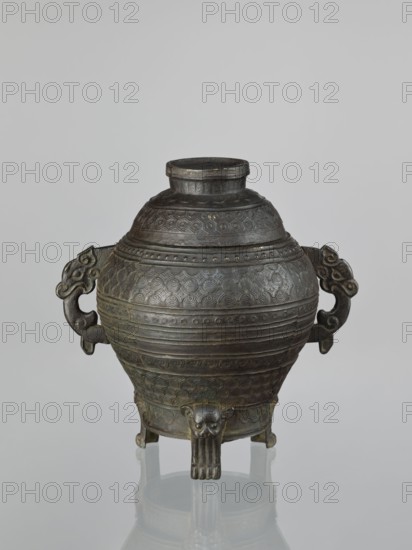 Dui-type vessel with a lid, censer/ritual/ceremonial vessel, Chinese, Qing dynasty 1644–1911, possibly 19th century, decorated with meander and leiwen ornaments and lion iconography, geometric ornament, bronze? material not specified, height 26.5 cm, width 26.5 cm, provenance unknown., A dark bronze vessel with a rounded body, a short neck, and a narrow rim sits on a reflective surface; the vessel has a pair of ornate handles shaped like stylized scrolls on opposite sides and rises on three short feet that terminate in paw-like forms, with decorative raised patterns covering its surface including bands of concentric swirls, geometric motifs, and horizontal ridges; the visible colors are dark brown, muted greenish-brown, and hints of black, set against a pale gray background and reflected faintly below.