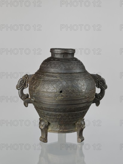 Dui-type vessel with a lid, censer; Chinese, Qing dynasty 1644–1911, 19th century, made in China. Ritual/ceremonial bronze vessel with leiwen thunder pattern, meander and geometric ornament, lion iconography. Height 26.5 cm; width 26.5 cm., Provenance unknown., An ornate round vessel sits against a pale gray background, made of dark bronze with areas of greenish patina and lighter brown highlights; it has a short cylindrical neck and a domed body decorated with repeating circular and geometric patterns in low relief, horizontal banding, and some small holes and surface irregularities; two stylized handles extend from opposite sides with curling, cloudlike shapes, and the vessel is supported by three short legs carved with animal-like faces and paw-shaped feet, the legs and lower body reflecting faintly on the glossy surface beneath.