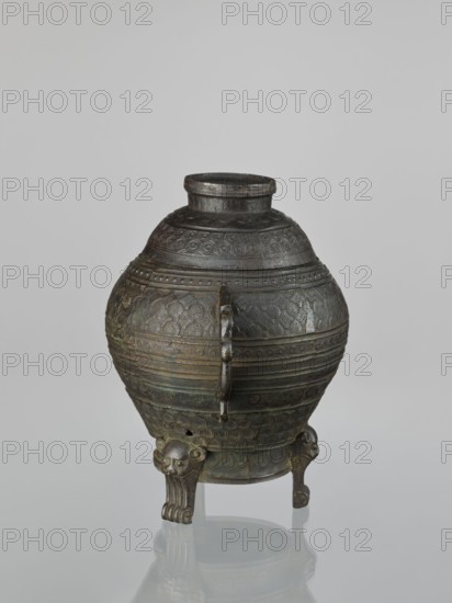 Dui-type vessel with a lid, censer, ceremonial/ritual vessel, made of bronze, featuring meander, leiwen, geometric ornament and lion iconography; Chinese, Qing dynasty 1644–1911, possibly 19th century; height 26.5 cm, width 26.5 cm., A rounded metal vessel with a narrow neck and a small opening sits on a reflective surface; the vessel is primarily dark bronze with areas of greenish patina and subtle brownish tones, and it is covered in repeating embossed geometric and circular patterns across its body. A vertical handle-like projection runs down the front, and the vessel rests on a group of ornate tripod legs each shaped with stylized animal faces and curved feet. The neck and shoulder display bands of concentric designs, and the overall finish shows aged metal textures and slight variations in color from dark brown to green.