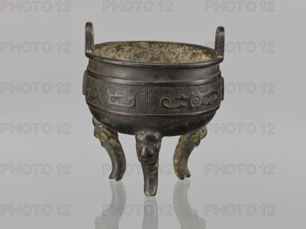 Ding incense burner, Chinese, ding ceremonial/ritual vessel with leiwen ornament, meander ornament, and dragons; Qing dynasty 1644–1911, 19th century, China; vessel; height 12.8 cm, diameter 10.5 cm., A small metal tripod vessel with a rounded bowl and two short vertical handles, standing on three curved legs that end in stylized animal-head forms; the bowl has a raised decorative band with geometric and curvilinear patterns encircling it, and the interior shows a mottled, mottled surface. The object displays dark brown and black tones with areas of greenish patina and hints of lighter brown and grey, set against a pale grey background, with reflections of the legs visible on the glossy surface beneath.