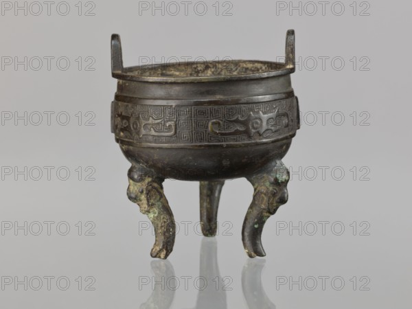 Ding incense burner, Chinese, ding vessel; ceremonial/ritual vessel with leiwen and meander ornament, dragons; Qing dynasty 1644–1911, 19th century, China; vessel; height 12.8 cm, diameter 10.5 cm., A small rounded metal vessel with two vertical handles projecting above its rim stands on three curved legs; the body shows an incised decorative band with geometric and stylized animal motifs around its upper portion, and the legs have sculpted animal-head forms with traces of green and brown patina; the metal surface displays shades of dark brown and bronze with areas of lighter greenish corrosion, and the vessel sits on a reflective pale gray surface against a neutral light gray background.