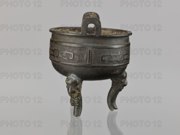 Ding incense burner, Chinese, ding vessel for ceremonial/ritual use, decorated with leiwen ornament, meander ornament, and dragons; Qing dynasty 1644–1911, 19th century, China; vessel; height 12.8 cm, diameter 10.5 cm, A small three-legged metal vessel with a rounded bowl and a raised rim sits against a pale gray background, showing a dark brown to black patina with areas of greenish corrosion; the bowl has a decorative band of incised geometric and curving motifs around its middle, and a flat looped handle rises from the rim at the back; each leg curves downward like an animal limb and ends in a rounded foot, with the legs and underside exhibiting patches of green and lighter brown where the surface has worn.