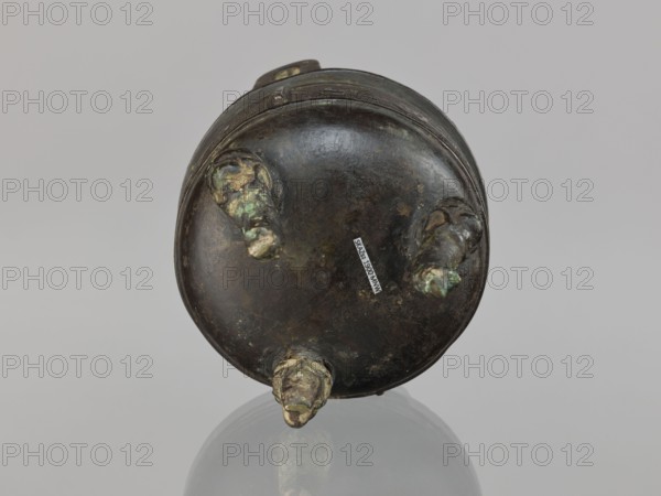 Ding incense burner, Chinese, ding vessel, ceremonial/ritual vessel, decorated with leiwen ornament, meander ornament, and dragons; Qing dynasty 1644–1911, 19th century, China; vessel; height 12.8 cm, diameter 10.5 cm., A round dark metal object with a patinated surface in shades of brown, dark gray, green, and some lighter green corrosion spots sits on a reflective surface, showing a circular base with a few short protruding stubs around its edge and a rounded rim, photographed against a plain light gray background.