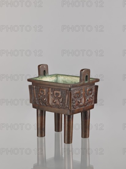 Fangding incense burner, Narozno Eckersdorf, Chinese art; fangding vessel; Chinese; ceremonial/ritual vessel; meander and leiwen ornament, taotie iconography; Qing dynasty 1644–1911; vessel standing on four cylindrical legs; 18th century, China; height 15.3 cm, width 13.1 cm, depth 10.3 cm., A bronze square vessel with a greenish interior patina sits on a reflective surface, supported by a group of four cylindrical legs; the vessel has a raised rim with two short vertical handles rising from opposite sides and a band of carved decorative patterns on its exterior, showing a darker brown bronze color with areas of lighter brown and greenish oxidation, and the reflected base and legs mirror these colors against a pale gray background.
