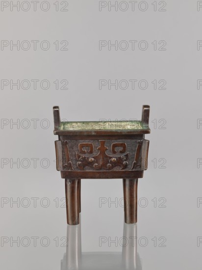 Fangding incense burner, Narozno Eckersdorf, Chinese art; fangding vessel; Chinese; ceremonial/ritual vessel; meander and leiwen ornament, taotie iconography; Qing dynasty 1644–1911; vessel standing on four cylindrical legs; 18th century; China; height 15.3 cm, width 13.1 cm, depth 10.3 cm., A small bronze vessel with a rectangular body and a shallow interior sits on three cylindrical legs; the body has a raised decorative band with stylized carved motifs and rounded bosses, and short vertical projections at two opposite corners above the rim. The interior surface shows a greenish patina while the exterior bronze ranges from dark brown to reddish-brown with subtle greenish corrosion in places, and the background is a neutral light gray.