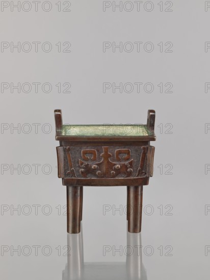 Fangding incense burner, Chinese, Qing dynasty 1644–1911, 18th century. Bronze vessel fangding standing on four cylindrical legs; ceremonial/ritual function. Decorations: meander and leiwen ornaments, taotie iconography. Dimensions: height 15.3 cm, width 13.1 cm, depth 10.3 cm., An object resembling a small rectangular bronze vessel with a greenish interior patina sits on three cylindrical legs; the exterior surface shows a dark brown bronze color with decorative raised patterns on the front panel, and the top corners have upward-facing short vertical projections.
