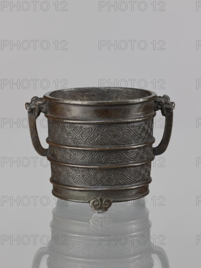Incense burner, revindication provenance — Chinese, Qing dynasty 1644–1911, 17th century, vessel with two handles; ritual/ceremonial censer; leiwen and meander ornament; height 12.6 cm, width 10.9 cm, diameter 8.2 cm., A small metal vessel with a dark brown and bronze patina sits centered against a light gray background, showing a cylindrical body with textured geometric patterns and several raised horizontal bands; it has a wide rim, a pair of curved handles with decorative terminals, and a small ornamental foot at the base, with a faint reflection visible beneath.