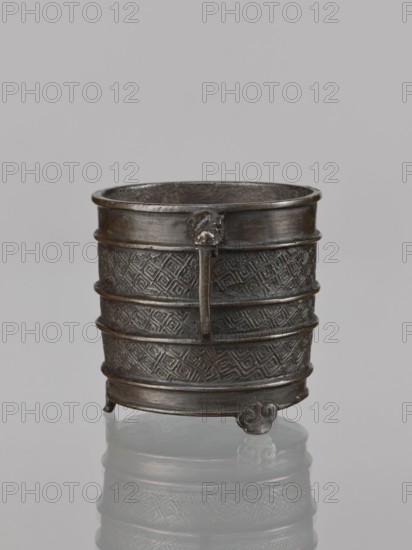 Incense burner, Chinese; censer with leiwen and meander ornament; ritual/ceremonial vessel with two handles; Qing dynasty 1644–1911, 17th century ; vessel; height 12.6 cm, width 10.9 cm, diameter 8.2 cm; China., A small cylindrical metal container stands on a reflective surface, showing a dark brown to black patina with areas of lighter brown highlights; the container has a slightly flared rim, several horizontal raised bands encircling its body, and a textured surface of repeating geometric diamond patterns between the bands; a vertical strap runs down the front and is attached near the rim with a small ornate element, and the base rests on three short decorative feet with curled shapes; the interior appears darker and rougher in texture, and the background is a smooth pale gray.