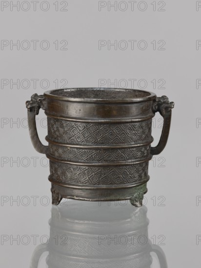 Incense burner, Chinese, Qing dynasty 1644–1911, 17th century, vessel with two handles; ritual/ceremonial censer with leiwen and meander ornament; height 12.6 cm, width 10.9 cm, diameter 8.2 cm., A small metal vessel with a dark brown and bronze patina sits against a pale gray background, featuring a cylindrical body decorated with a geometric diamond pattern banding and horizontal raised rims; two curved handles attach near the rim with stylized animal head mounts, and the base rests on small molded feet that show hints of greenish oxidation.