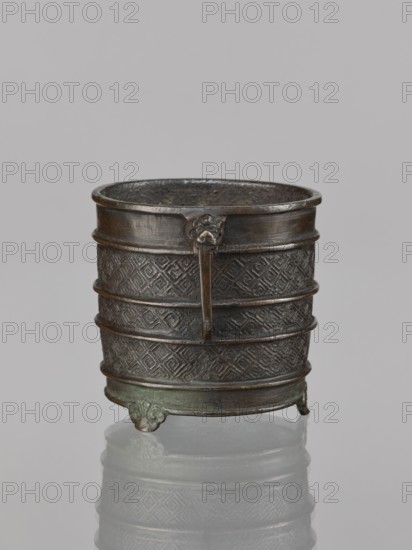Incense burner, Chinese, Qing dynasty 1644–1911, 17th century, vessel with two handles, ritual/ceremonial censer; decorated with leiwen meander ornament. Materials: vessel; dimensions: height 12.6 cm, width 10.9 cm, diameter 8.2 cm., A cylindrical metal vessel with a dark brown and greenish patina sits on a reflective surface, showing a textured banded body with raised horizontal ribs and repeating geometric patterns; a narrow vertical handle with a small decorative element at its top projects from the rim, and the vessel stands on a group of small decorative feet, with visible interior surface texture and subtle variations of dark brown, bronze, and green tones.