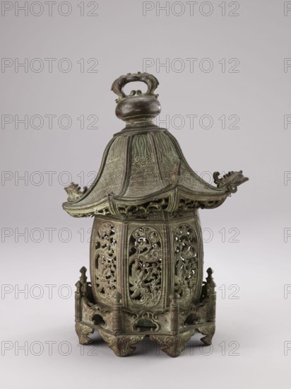 Lantern, gift; Genius, Mieczyslaw 1853–1920. Chinese, Qing dynasty 1644–1911, 19th century. Materials/decoration: fish, arbors, plant ornament, garden arbors. Height: 35.0 cm., An ornate metal object with a pagoda-like roof and a rounded finial handle sits against a plain light background; the piece shows a weathered greenish-brown patina with darker brown accents, and the roof has textured panels and upward-curving eaves ending in stylized creature heads. The body beneath the roof is an openwork cylindrical form with intricate scrolling and foliate cutouts visible through several vertical panels, and the lower section rests on a decorative base with pierced gallery-like railings, small finials, and carved bracket feet all sharing the same aged green and brown coloration.