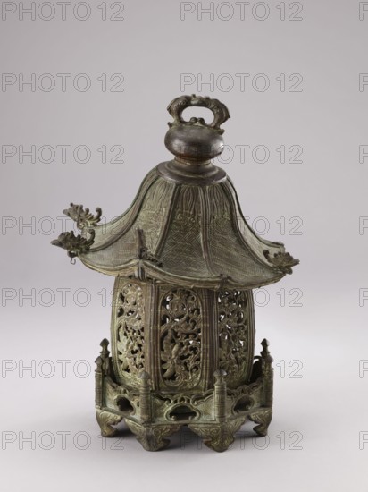 Lantern, gift; Genius, Mieczyslaw 1853–1920. Chinese, Qing dynasty 1644–1911. Decorated with fish, arbors, plants and garden arbors. 19th century, China. Height 35.0 cm., An ornate metal lantern-like object with a greenish-brown patina, featuring a domed, pagoda-style roof with a round finial and a decorative loop handle on top; the roof has textured, woven-patterned panels and upturned eaves with small dragon or creature head ornaments at the corners. Below the roof is a cylindrical body with openwork panels carved with scrolling floral and foliate motifs, surrounded by vertical ribs. The base is a raised platform with a low balustrade of small posts and pierced arches, standing on carved feet with curving shapes. The visible colors are green, brown, and hints of darker bronze.