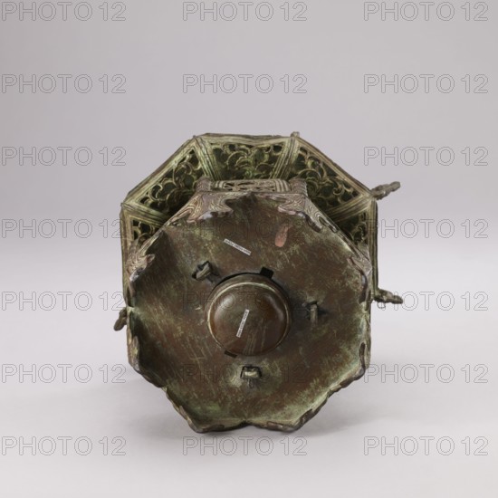 Lantern, Qing dynasty 1644–1911, 19th century, China; materials/technique: not specified; decorated with fish, arbors, and plant/garden arbor ornament; height 35.0 cm., A metallic object with a worn, patinated surface sits against a light background, showing shades of green, brown, and hints of reddish-brown; the object has an octagonal base with scalloped edges and decorative raised elements, and a central rounded protrusion surrounded by small bracket-like features; the body above the base displays openwork panels with intricate scrolling patterns and a darker green patina, and there are decorative handles or extensions visible on the sides in similar green and brown tones.