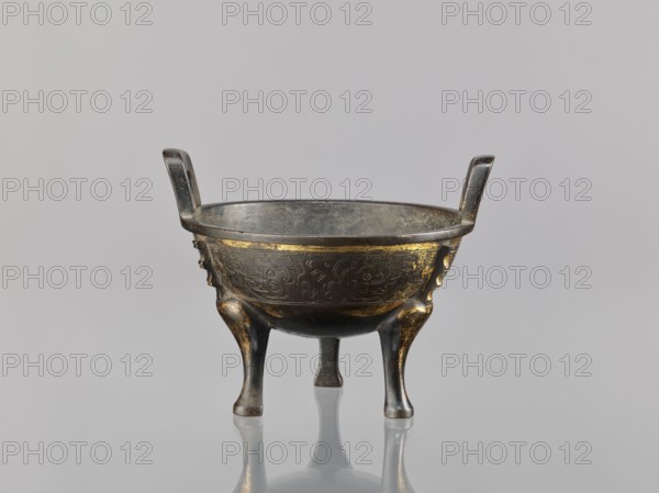 Ding incense burner, Chinese; decorated with fantasy beasts, meander and leiwen ornament; Qing dynasty 1644–1911, 19th century, ceremonial/ritual vessel censer; height 12.6 cm, width 16 cm, diameter 14.5 cm., An aged metallic vessel with a round bowl supported by three curved legs and two upright handles, showing a dark gray and bronze patina with areas of gold-colored accents and worn highlights; the bowl's exterior surface has subtle raised decorative patterns and worn gilding, and the interior appears darker and more matte; the object is photographed against a smooth light gray background with a faint reflective surface below producing a soft mirror image.