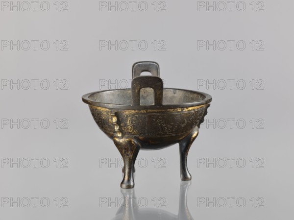 Ding incense burner, Chinese, Qing dynasty 1644–1911, 19th century, bronze vessel censer used for ritual/ceremonial purposes, decorated with fantasy beasts, meander and leiwen ornament, height 12.6 cm, width 16 cm, diameter 14.5 cm., A small three-legged metal vessel sits centered against a plain light gray background, showing a rounded bowl with a thick rim and two vertical rectangular handles rising from opposite sides; the vessel has a dark bronze appearance with patches of gold-toned highlights and worn areas, and decorative raised patterns around the bowl's exterior; the three curved legs taper toward rounded feet and show the same dark bronze and gold coloration, with a shiny reflective surface beneath the vessel that mirrors its legs and underside.