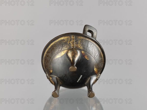 Ding incense burner, Chinese, beasts fantasy, meander ornament, leiwen ornament, Qing dynasty 1644–1911, ceremonial/ritual vessel, 19th century, China; vessel/censer; height 12.6 cm, width 16 cm, diameter 14.5 cm., A rounded metal vessel with a short handle and three curved legs is shown from a low frontal angle, revealing an underside with a domed bottom and a slightly flared rim. The surface shows areas of worn metallic sheen and patterned decorative engraving along the upper band. The legs and central rounded knob exhibit patches of brighter metallic highlights and darker patina, and there is a small rectangular label attached to the lower side. The visible colors include dark brown, black, gold, and hints of gray.